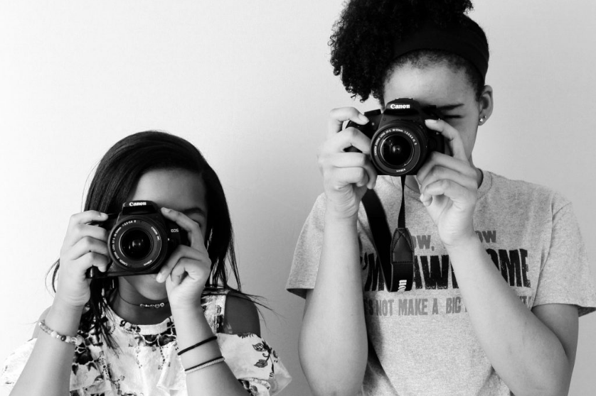A black and white photo. Two young Black girls look through the viewfinder of Canon DSLRs, their faces obscured. They point their cameras at the viewers head-on.